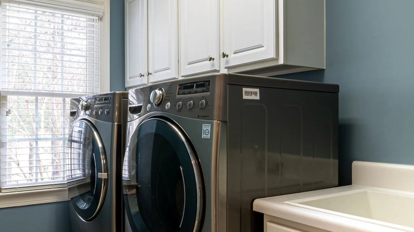 Un sèche-linge gris à hublot rond, posé à côté d'un lave-linge sous des meubles blancs dans une buanderie